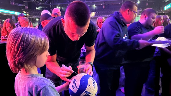 Der Kanadier Cole Ketrzynski schreibt ein Autogramm auf den Volleyball eines kleinen Fans. © NDR Niedersachsen Foto: Marie Schiller