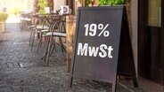 Auf einer Tafel vor einem Gastronomiebetrieb steht ein Schild mit der Aufschrift 19 Prozent Mehrwertsteuer. © picture alliance / CHROMORANGE | CHROMORANGE 