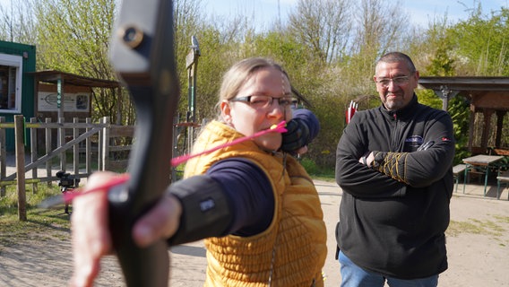 Kursteilnehmerin zielt mit Pfeil und Bogen, Trainer Frank Schrader beobachtet sie dabei auf seinem Trainingplatz in Hannover. © NDR Foto: Marcel Seemann