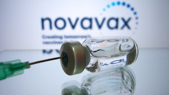Das Bild zeigt eine Nadel und eine Impfstoff-Ampulle vor einem Novavax-Hintergrund. © picture-allaince Foto: Sven Simon