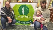 Die Gründer der Dorfschule in Oberndorf posieren mit ihrem Kind auf dem Arm vor dem Logo der Schule. © NDR Foto: Birgit Langhammer