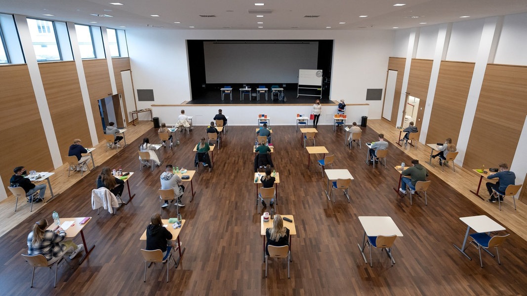 Schülerinnen und Schüler eines Gymnasiums sitzen vor Beginn ihrer schriftlichen Prüfung in einer Aula an ihren Plätzen.