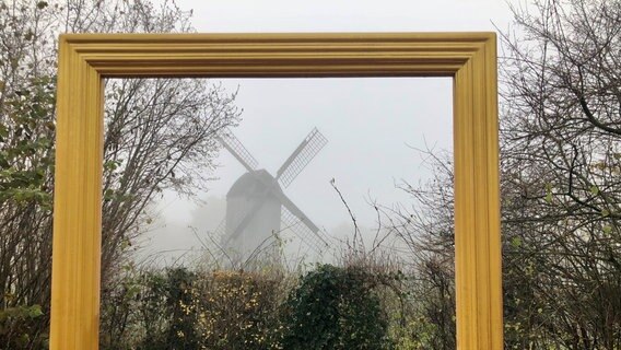 Durch einen Bilderrahmen um Vordergrund ist eine Windmühle im Nebel zu sehen. © NDR Foto: Thomas Schwierzi