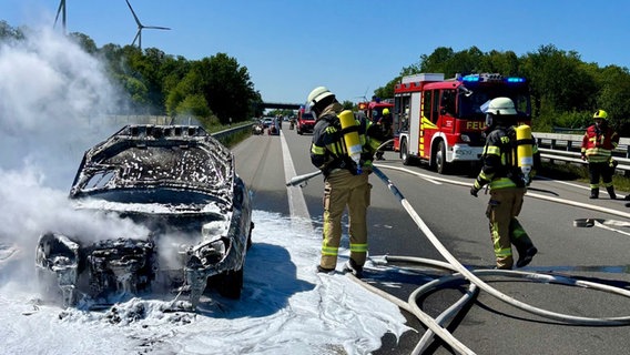 Feuerwehrleute stehen vor einem ausgebrannten, noch qualmenden Auto auf einer Autobahn. © Feuerwehr Riepe Foto: Dorian Kersten