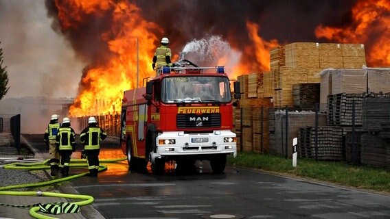 Feuerwehrleute stehen mit einem Einsatzwagen vor einem Haufen brennender Paletten auf einem Firmengelände. © NonstopNews 