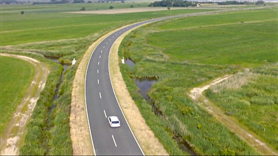 Auf einer Straße fährt ein Auto zwischen grünen Feldern. © NDR 