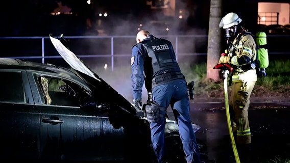 Ein Polizist untersucht ein ausgebranntes Auto in Oldenburg, eine Person in Feuerwehrmontur steht daneben. © NonstopNews 