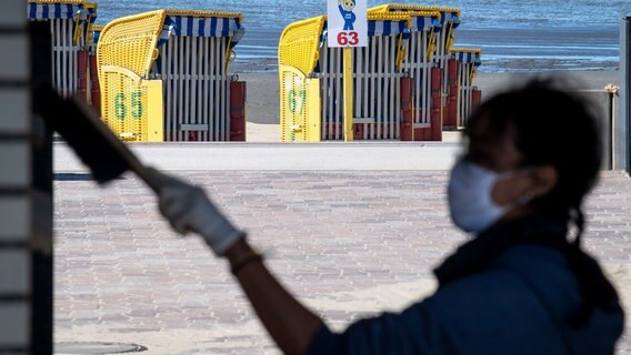Eine Frau mit Atemschutzmaske bürstet die Fassade eines Gebäudes vor einem Strand ab. © dpa-Bildfunk Foto: Sina Schuldt