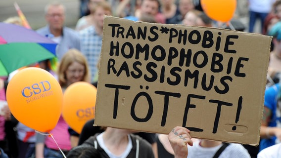 Teilnehmer des Cristopher Street Days in Oldenburg halten ein Schild mit der Aufschrift "Trans*Phobie, Homophobie, Rassismus tötet" in die Höhe. © picture alliance/dpa Foto: Ingo Wagner
