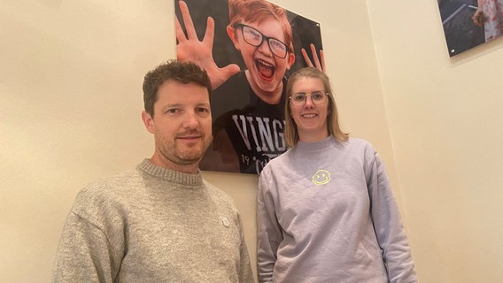 Man sieht ein Paar vor dem Bild ihres Sohnes, der eine fröhliche Grimasse macht Man sieht ein Paar vor dem Bild ihres Sohnes, der eine fröhliche Grimasse macht © NDR Foto: Fritzi Blömer