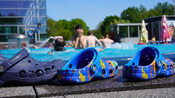 Neben einem Freibad-Becken stehen Badelatschen, im Hintergrund sind Kinder im Becken zu sehen. © NDR Foto: Lotti Höfer