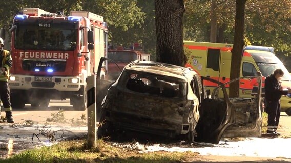 Nach einem Unfall steht ein ausgebranntes Auto an einem Baum, im Hintergrund sind Rettungskräfte zu sehen. © NonstopNews 