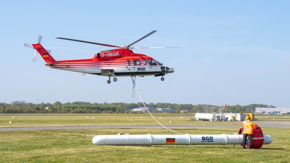 Ein rot-weißer Hubschrauber hebt ab und transportiert an einem langen Seil eine Forschungssonde, um das Grundwasser in Niedersachsen zu untersuchen. © dpa-Bildfunk Foto: Melissa Erichsen