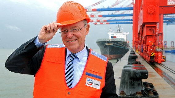 Stephan Weil (SPD) mit Schutzhelm und Warnweste mit der Aufschrift "Eurogate" am JadeWeserPort. Er lächelt und hält eine Hand grüßend an den Schutzhelm. © picture alliance Foto: Ingo Wagner