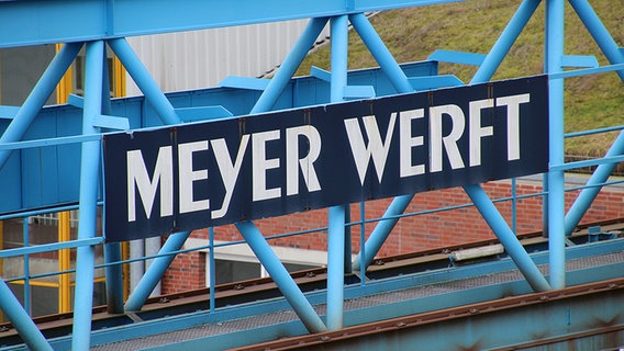 An einem blauen Metallgerüst hängt ein Schild mit der Aufschrift "Meyer Werft". © NDR Foto: Oliver Gressieker