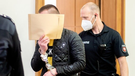 Der Angeklagte sitzt vor Prozessbeginn mit Handfesseln in einen Saal im Landgericht und bedeckt sein Gesicht mit einem Briefumschlag. Der junge Mann soll im März 2021 in Emstek die drei Monate alte Tochter seiner damaligen Lebensgefährtin durch Schütteln und Schlagen auf eine harte Oberfläche getötet haben. © dpa-Bildfunk Foto: Hauke-Christian Dittrich