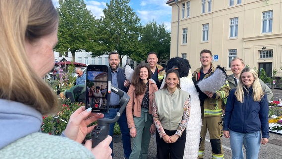 Eine Frau filmt eine Gruppe von Menschen, einer von ihnen in einem Pinguinkostüm, auf dem Wochenmarkt in Oldenburg. © NDR Foto: Nadia Yaqub