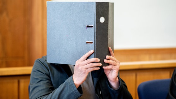 Ein Angeklagter in einem Mordfall sitzt hält sich einen Aktenordner vor das Gesicht. © Hauke-Christian Dittrich/dpa Foto: Hauke-Christian Dittrich