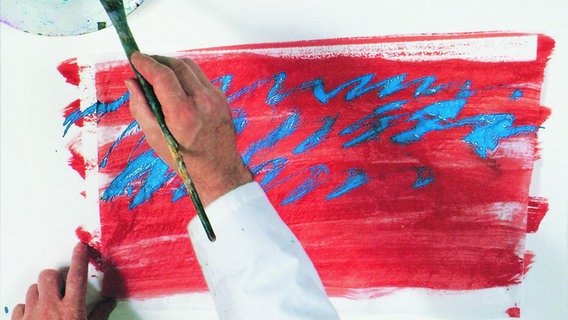 Die Hand eines Mannes hält einen Pinsel, auf einer Arbeitsfläche liegt ein rotes Bild mit blauen Wellenlinien. © Eric Carle Studio / Gerstenberg Verlag 