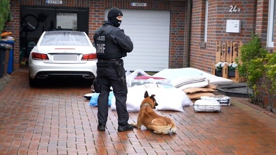 Ein Beamter der Polizei steht vermummt mit einem Spürhund vor einem Haus in Lohne, bei dem eine Razzia druchgeführt wurde. © Nord-West-Media TV 