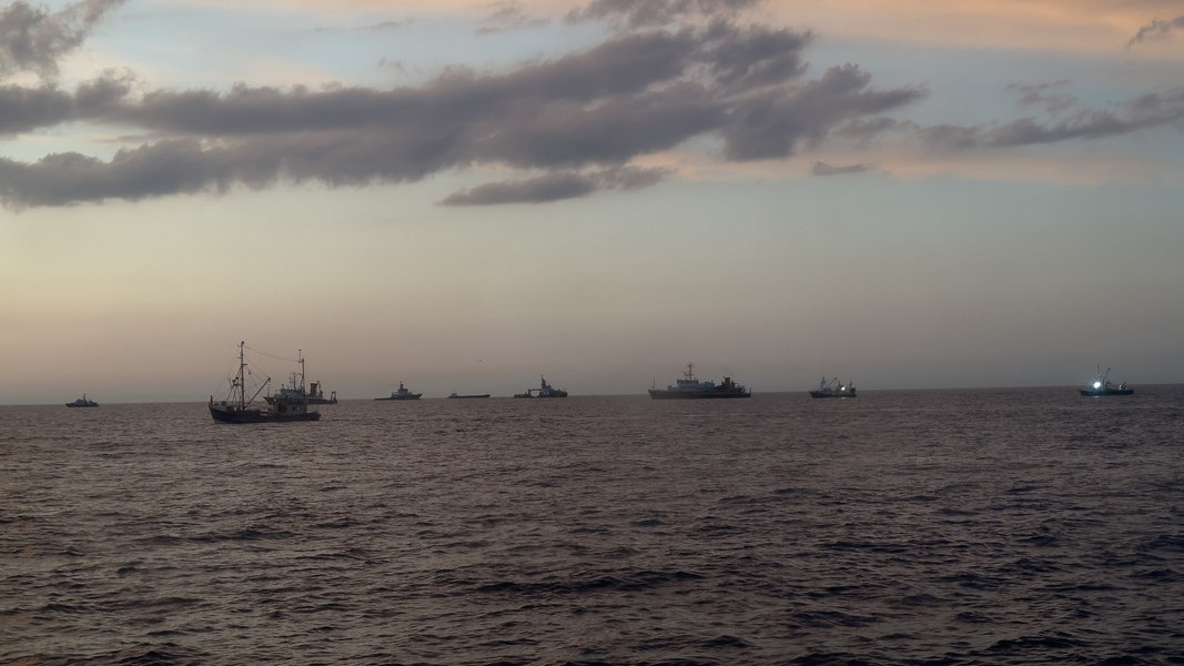 Mehrere Schiffe und Boote schwimmen in der Abenddämmerung auf dem Wasser, einige von ihnen haben Scheinwerfer angeschaltet.