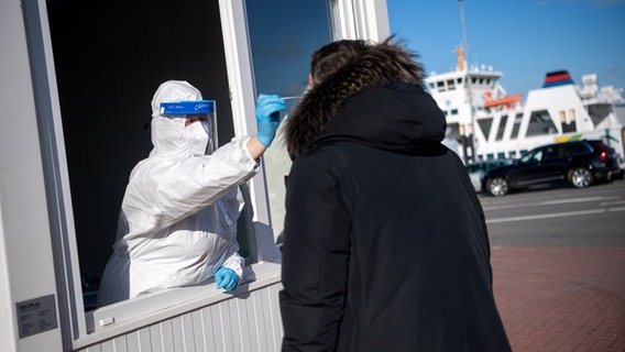 Eine Frau in weißem Schutzanzug, mit Maske und Visier steht in einem Container am Fähranleger Norddeich und führt bei einer Person einen Corona-Test durch. © dpa-Bildfunk Foto: Sina Schuldt