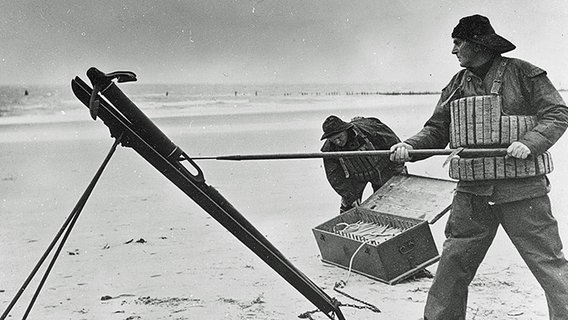 Eine historische Fotografie zeigt zwei Männer, die am Strand eine Leuchtrakete abfeuern. © DGzRS/Die Seenotretter Foto: DGzRS