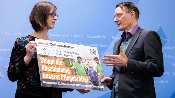 Eine Frau übergibt einem Mann ein großes Plakat mit der Aufschrift: "Stoppt die Abschiebung unserer Pflegekräfte!" © picture alliance/dpa Foto: Carsten Koall