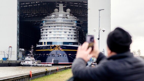 Das Kreuzfahrtschiff ·Disney Wish· wird von Schleppern aus dem Baudock der Meyer Werft gezogen und spiegelt sich dabei in einer großen Pfütze. © picture alliance/dpa Foto: Hauke-Christian Dittrich