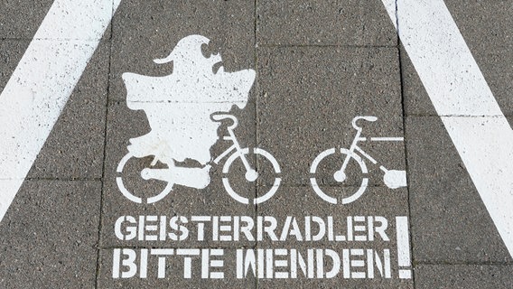 Auf einem Radweg ist mit weißer Farbe ein Warndreieck mit zwei sich entgegenkommenden Rädern abgebildet. Auf einem Rad fährt ein Gespenst. Der Text darunter sagt: Geisterradler, bitte wenden! © picture alliance | Christian Ohde Foto: Christian Ohde