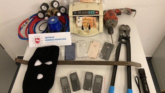 Von der Polizei sichergestellte Beweise wie ein Seitenschneider, Telefone, Gasschläuche, Geld und ein Bohrmaschine liegen auf einem Tisch. © Polizeidirektion Osnabrück 