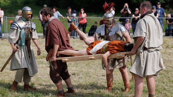 Als Römer verkleidete Schauspieler machen mit bei einem Schaukampf und tragen einen "Verletzt" auf einer Trage davon. © dpa Foto: Friso Gentsch