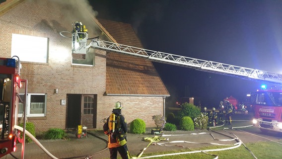 Einsatzkräfte der Feuerwehr stehen auf einer Drehleiter, aus dem Zimmer eines Hauses kommt Rauch nach draußen. Wasserschläuche liegen auf dem Boden. © Feuerwehr SG Nordhümmling 