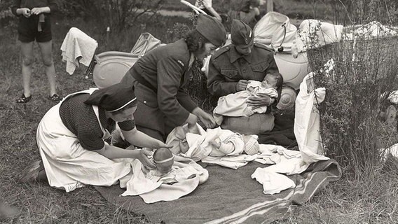 Eine Schwarz-Weiß-Aufnahme von drei Kleinkindern, die von drei Erwachsenen umsorgt werden. © Library and Archives Canada 