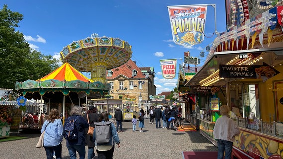 Personen gehen über eine Straße, rechts eine Pommesbude, im Hintergrund ein Kettenkarussel. © NDR Foto: Patricia Averesch