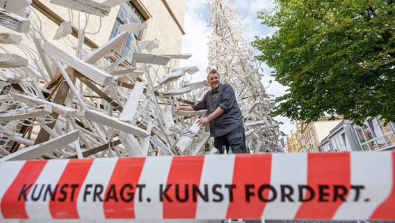 Der Künstler Volker-Johannes Trieb steht vor seiner Kunstinstallation "forx.pitchforks for peace" anlässlich 375 Jahre Westfälischer Frieden. Im Vordergrund ein Flatterband mit dem Aufdruck "Kunst fragt. Kunst fordert." © dpa-Bildfunk Foto: Friso Gentsch