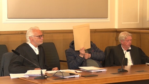Ein Angeklagter sitzt auf einer Bank, neben ihm seine Verteidiger. Er verdeckt sein Gesicht mit einem Briefumschlag. © Nord-West-Media TV 