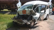 Ein demolierter Schulbus steht auf einer Straße, im Hintergrund begutachten Arbeiter einen Zug. © Nord-West-Media TV 