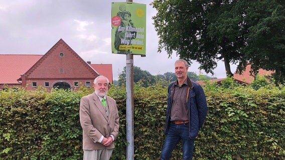 Wahlhelfer Herbert Seidel (Grüne) und Bürgermeister Reinhard Kurlemann (CDU) vor einem Wahlplakt der Grünen, das an einer Straßenlaterne befestigt ist. © NDR Foto: Hedwig Ahrens