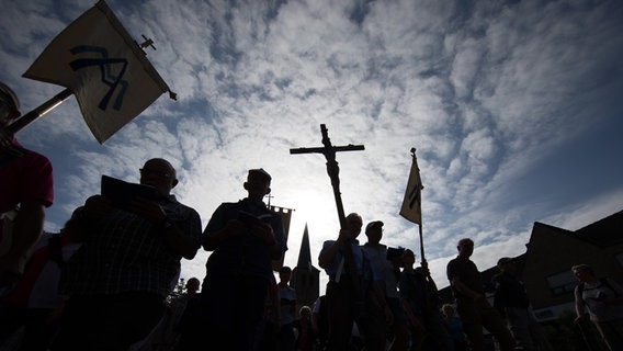 Pilger der 166. Telgter Wallfahrt gehen im Gegenlicht auf einer Straße und halten ein Kreuz. © dpa-Bildfunk Foto: Friso Gentsch