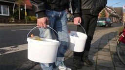 Zwei Schlachthofarbeiter tragen in weißen Eimern ihren Proviant. Zwei Schlachthofarbeiter tragen in weißen Eimern ihren Proviant. © NDR Foto: Hedwig Ahrens