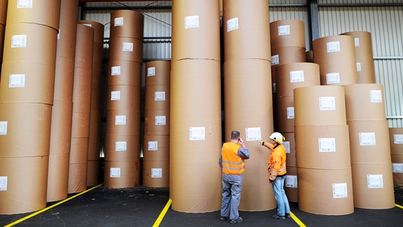 Zwei Arbeiter mit orangen Warnwesten stehen in einer Papierfabrik vor Papierrollen. © picture alliance/dpa Foto: Jonas Güttler