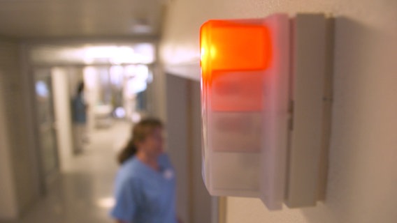 Eine Lampe über einem Krankenzimmer leuchtet rot, eine Pflegerin geht in Richtung Zimmer. © NDR Foto: Screenshot