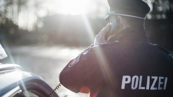 Policjant w mundurze stoi obok radiowozu pod światło i trzyma w ręku słuchawkę telefoniczną.  © NTR Zdjęcie: Julius Matuchik