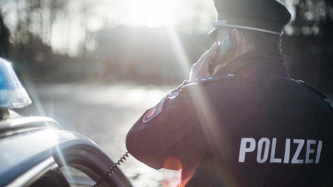 Ein Polizist in Uniform steht bei Gegenlicht neben einem Polizeiauto und hält einen Telefonhörer in der Hand.