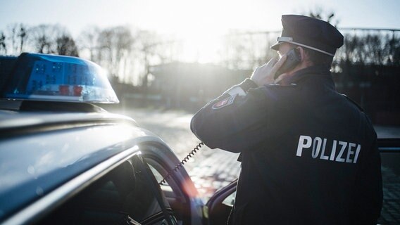 Ein Polizist in Uniform steht neben einem Polizeiauto und hält einen Telefonhörer in der Hand. © NDR Foto: Julius Matuschik