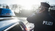 Ein Polizist in Uniform steht neben einem Polizeiauto und hält einen Telefonhörer in der Hand. © NDR Foto: Julius Matuschik