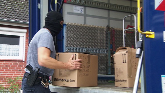 Ein Polizist mit Sturmhaube lädt Kartons mit beschlagnahmten Beweismittel in einen Lkw. © NDR 