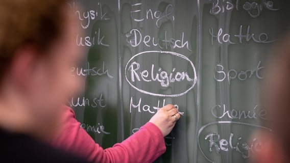 Eine Lehrerin schreibt mit Kreide einen Stundenplan mit Religionsunterricht an eine Tafel. © picture alliance/dpa Foto: Friso Gentsch