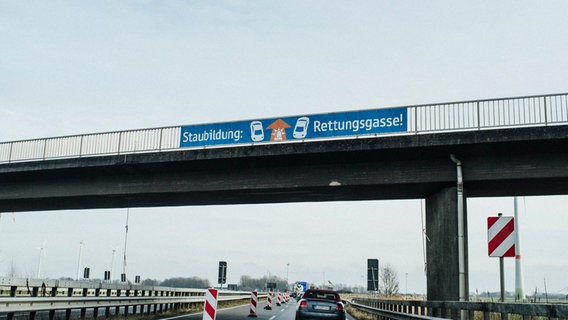 An dem Geländer einer Autobahnbrücke hängt ein Hinweisschild mit der Aufschrift Staubildung: Rettungsgasse. © NDR Foto: Julius Matuschik